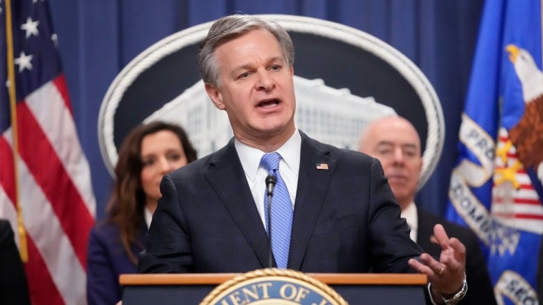 A man in a suit and tie is shown speaking at a podium with a seal on it. Behind him, a man and a woman stand.