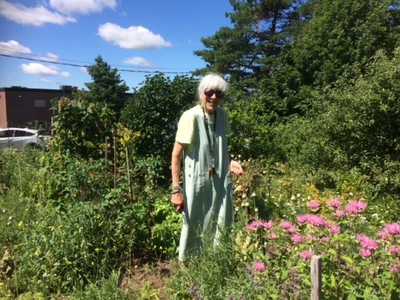 a woman stands in a  garden wuill tall palnts and trees. 