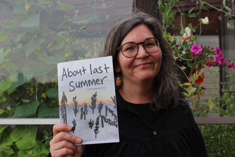 A woman standing in front of a greenhouse holds up a paper pamphlet. 