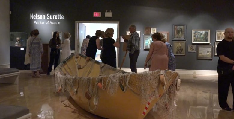 A large boat is seen on display in the middle of an art gallery. People mill about and paitnings can be seen on the walls.