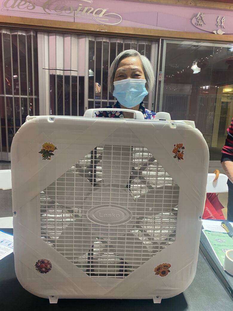 An older woman with a blue face mask stands behind her DIY air cleaner, made of a box fan taped to an air filter. The fan is decorated with a few stickers of flowers.