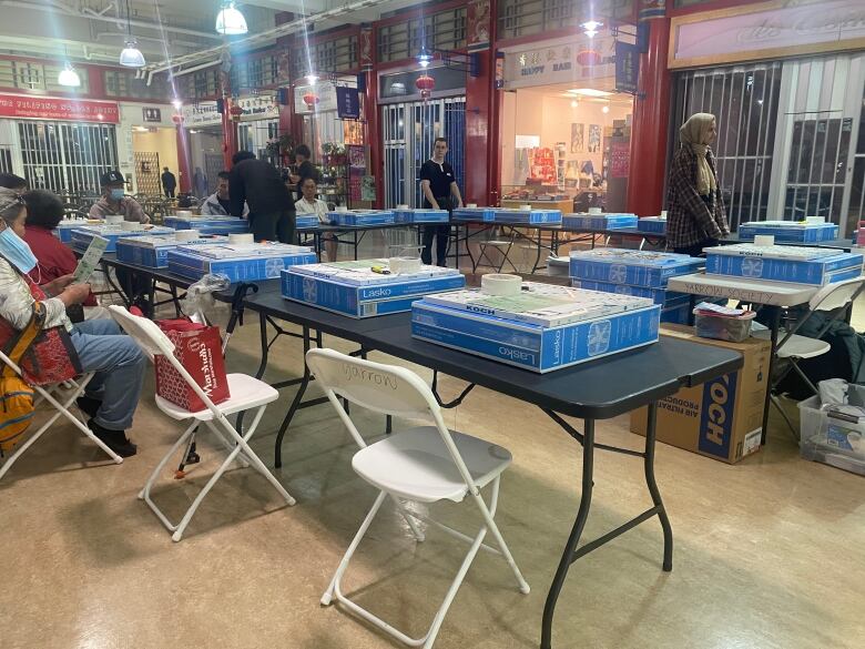 Fold-out tables are arranged in a U-shape in a mall. Around the tables a few people sit in chairs and wait for instruction. Large boxes of supplies sit in front of them on the tables.