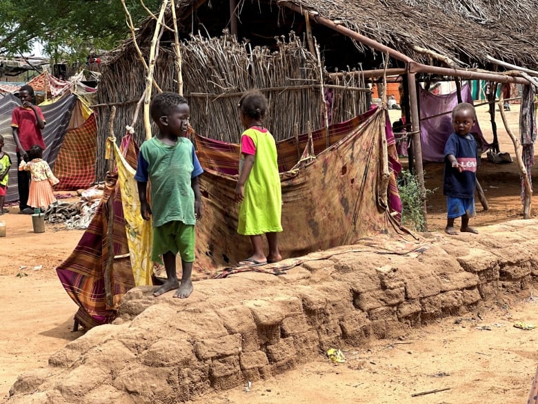 Small children are seen walking around makeshift huts on the dirt-covered ground.