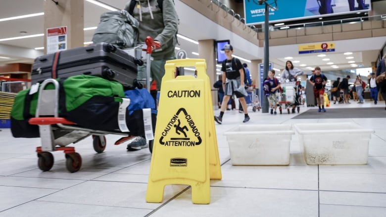 A man pushing a kart with luggage stacked on it through an airport navigating around a yellow 'caution' sign.