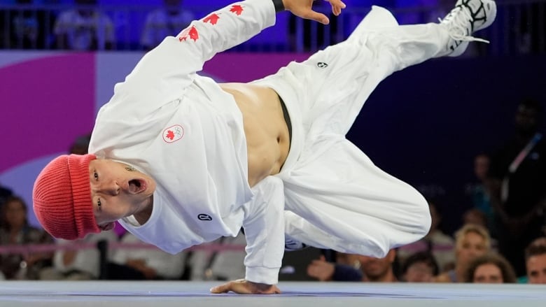 A man athletically poses in a breaking 'airchair' freeze. He holds himself vertically with one hand on the ground, as the rest of his body balances on his elbow which is jammed into his side.