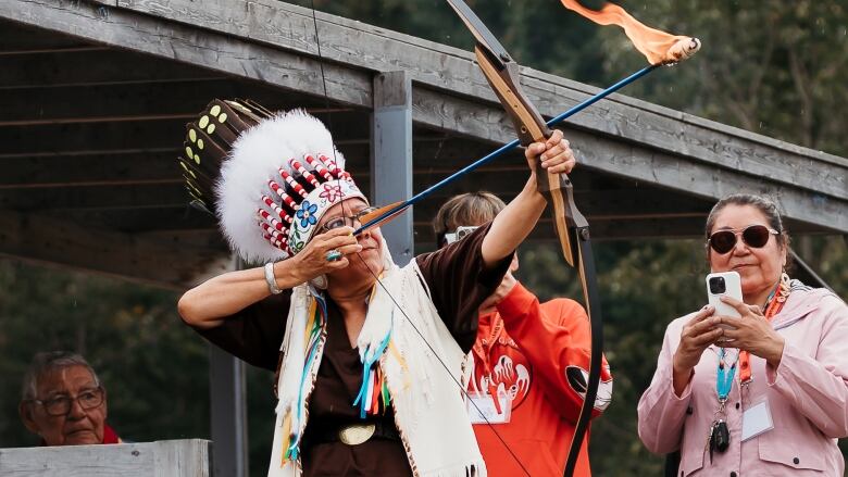 A woman in a headdress and ribbon skirt prepares to shoot a flaming arrow with a bow.