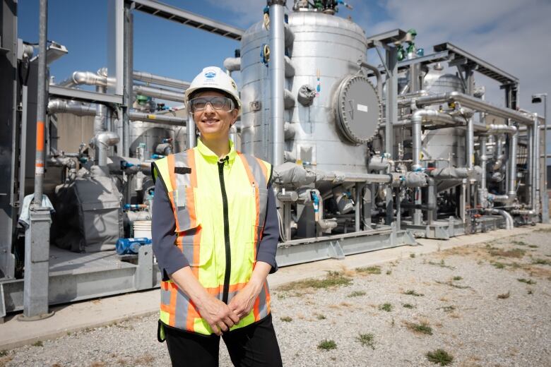 A woman in safety gear stands in front of a huge machine made of silver tubes and tanks