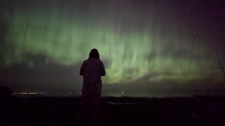 A silhouetted figure stands before a sky lit up in green and yellow colours. 