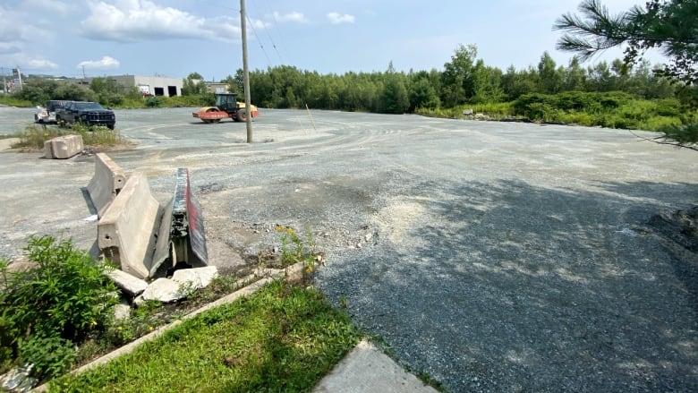 A large gravel piece of land is seen surrounded by trees, with a black truck parked on it and a piece of construction equipment