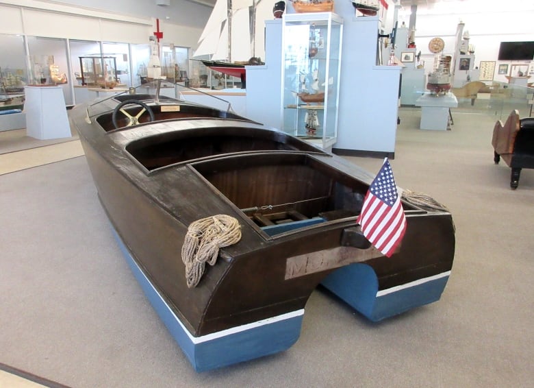 An American flag sits on the back of a wooden boat. The boat is sitting on the floor of a museum.