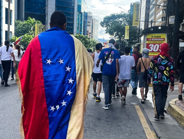 People with their faces turn walk in a street. One is draped in a Venezuelan flag.