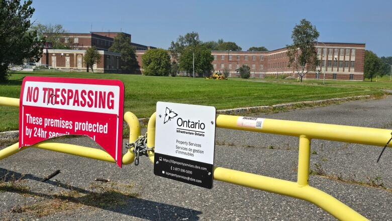 A yellow gate with signs reading no trespassing and Infrastructure Ontario, with a large brick building in the background.