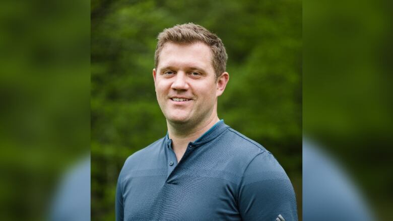 A person with short, light hair in a blue shirt smiles, green trees are in the background.