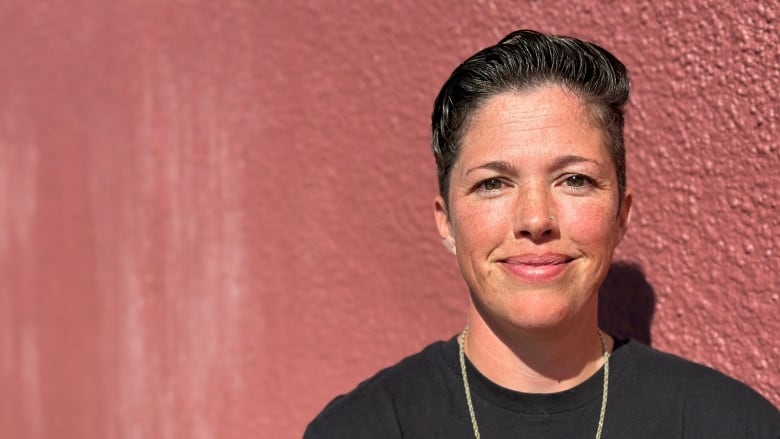 A woman with short dark hair and a black t-shirt stands in front of a red wall.