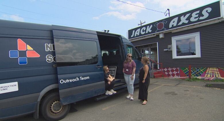 Three women stand next to blue van that says outreach team, outside of a building with a sign that says Jack Axes.