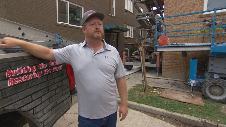 A man wearing a baseball cap leans against his truck at a construction site. 