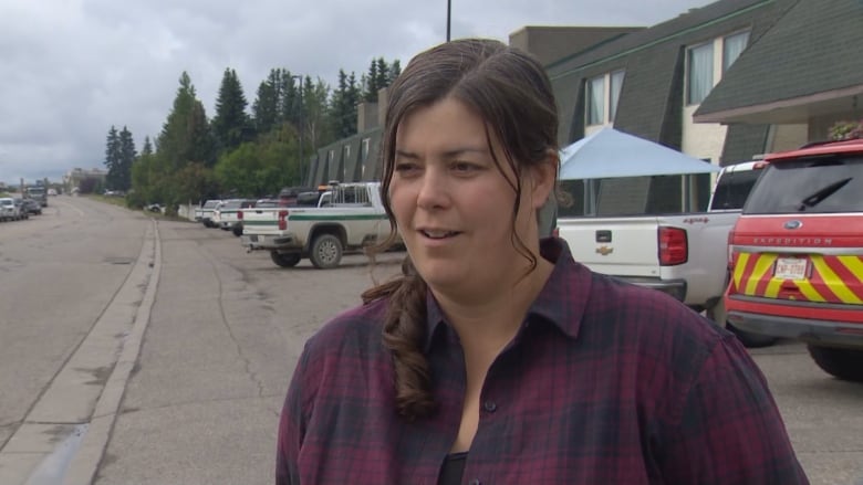 A white woman, with brown hair in a ponytail, is wearing a navy-and-maroon flannel shirt. She is standing outside, in front of multiple pickup trucks.