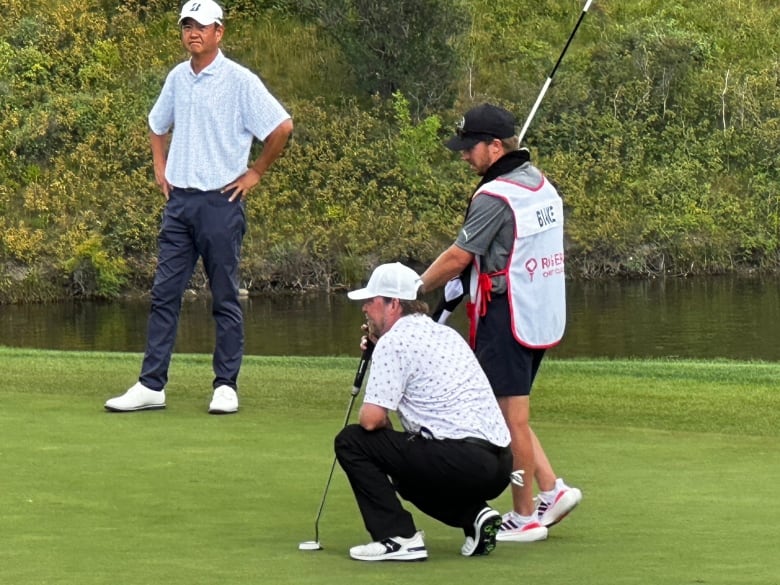 A man with a light blue shirt and navy pants and a white cap watches while a man with a white spotted shirt and black pants kneels holding a putter. A man wearing a grey shirt and black shorts stands behind the kneeling man.