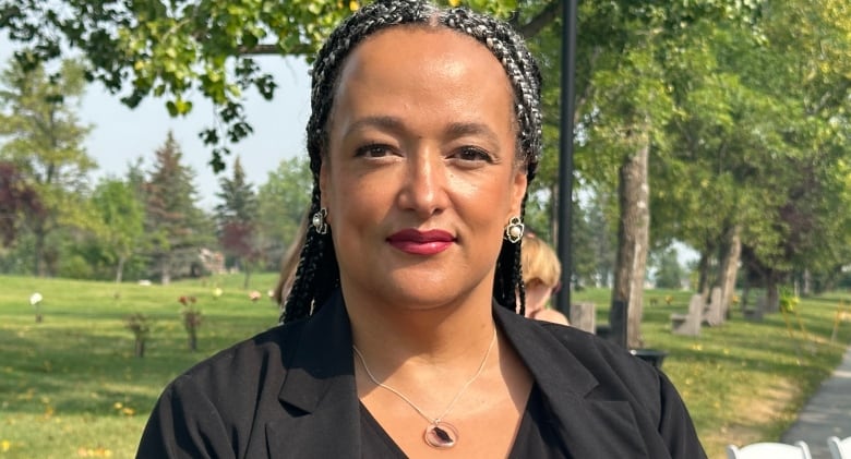 A woman with braided hair, wearing a black blouse and blazer, looks to the camera.