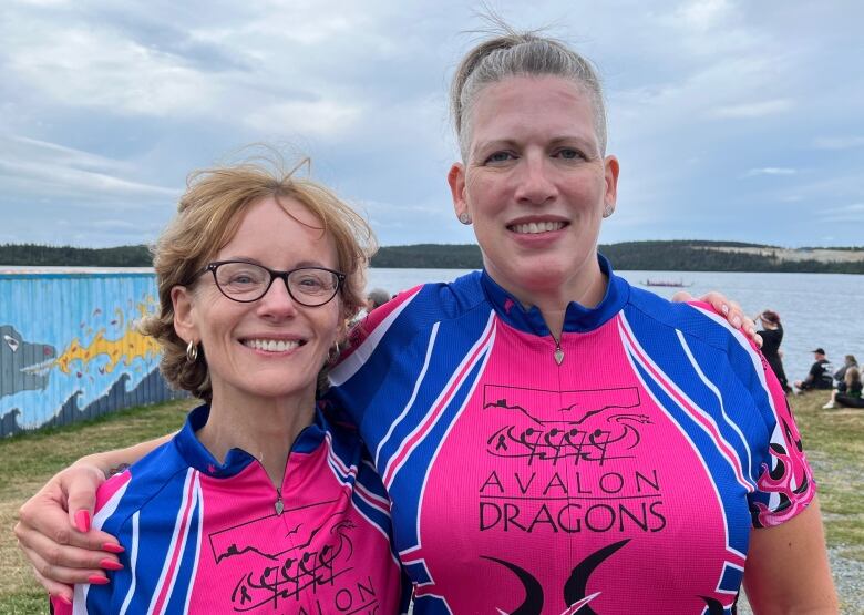 Two women stand next to each other and smile. The woman on the right has her arm around the other woman's shoulder. Both wear wetsuits.