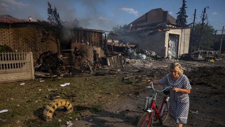 An older woman with a bicycle is shown near residential structures that are charred by fire or damaged.