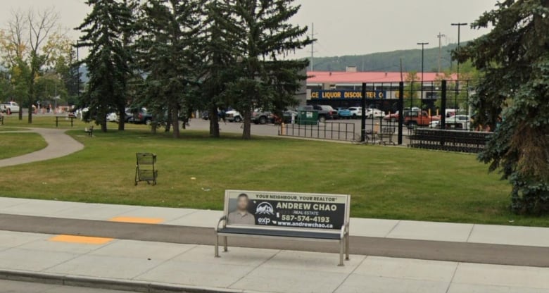 A bus stop bench is seen in the foreground and behind it is a small park with a pathway winding through it to shops in the distance.