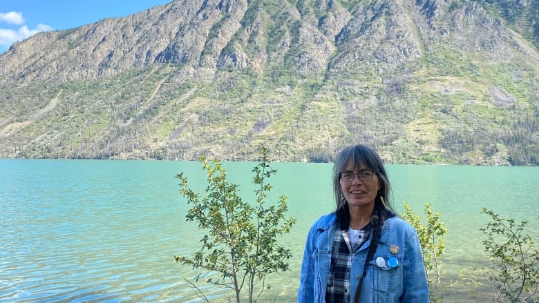 A woman in a denim jacket and flannel shirt stands in front of clear blue water and a mountain in the back drop. 