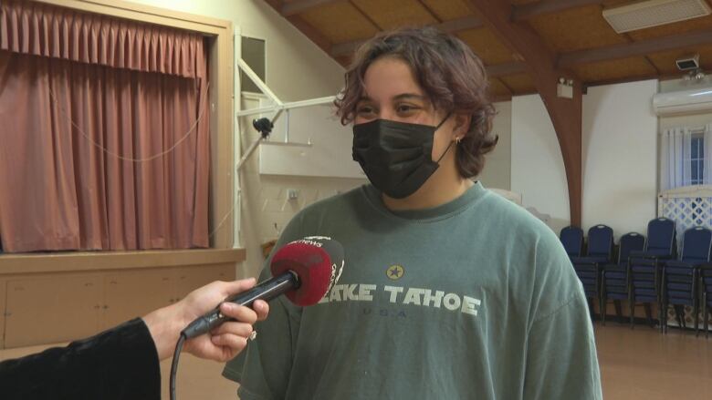 A man with medium length wavy hair speak into a microphone. He is wearing a black face mask, his ears are pierced and his shirt is blue. He is standing inside a community building.