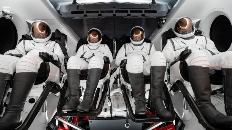 Four people dressed in white spacesuits with black boots sit in a spacecraft with their sun visors down.