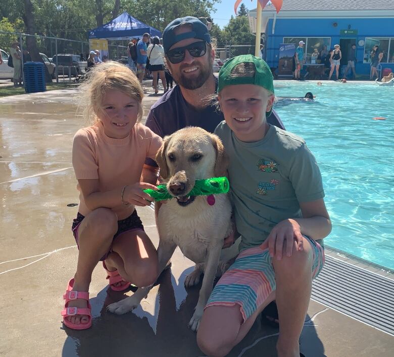 Two children flank a man and a dog while posing for a photo at an outdoor pool.