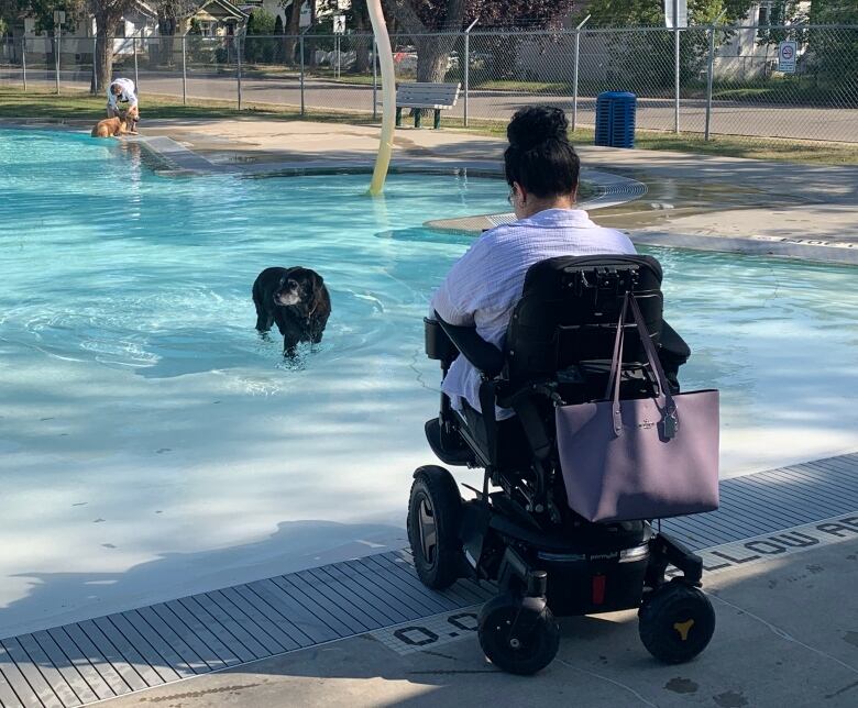 A woman in a motorized wheelchair watches a dog swim in an outdoor pool.