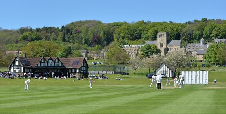 A group of people playing cricket on a lawn with large, old buildings in the background.