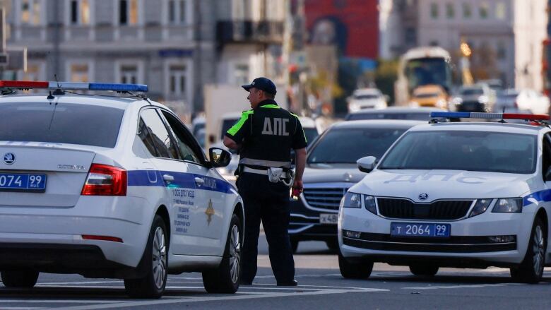 A police officer wearing a baseball cap is shown in a busy street between vehicles.