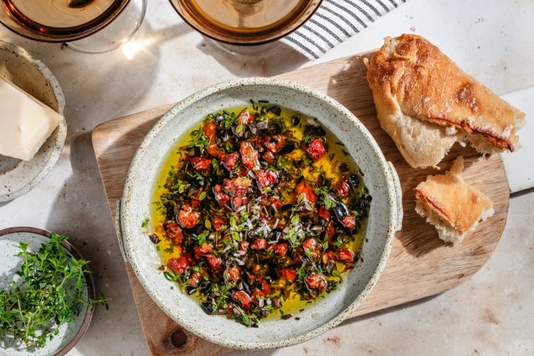 A serving bowl filled with a mediterranean dipping oil, on a wooden platter, next to a crusty loaf of torn bread.