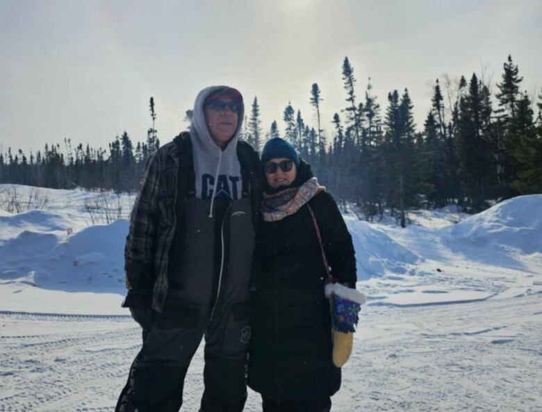 A older Indigenous couple stand on a cold winter day surrounded by snow looking at the camera.