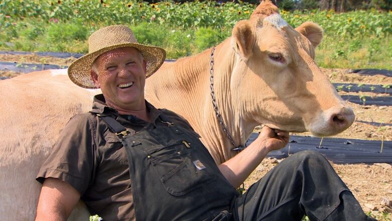 Man wearing a straw hat and overalls sits next to golden coloured cow in field with sun flowers in the background.