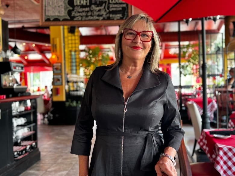 A woman with shoulder length blonde hair wearing red glasses and red lipstick is shown smiling in her restaurant, which is also decorated in red.