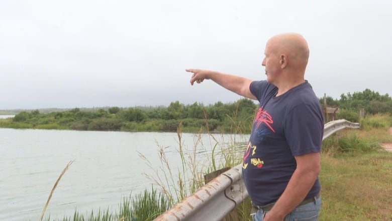 A man stands behind a guard rail pointing to a large pond. He is in a blue t-shirt. 