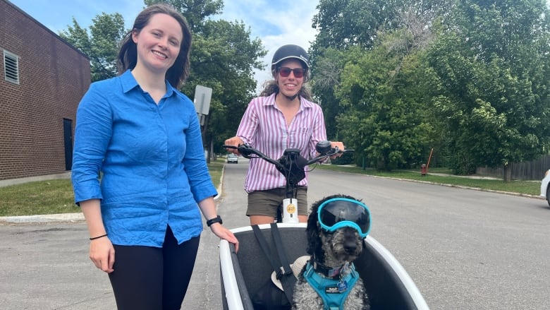 A woman in a blue shirt is standing next to a woman on a cargo bike, with a dog in the front compartment.