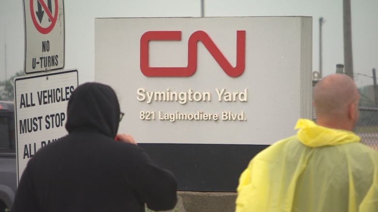 Rail workers stand in the rain outside of a sign that says CN Symington Yard