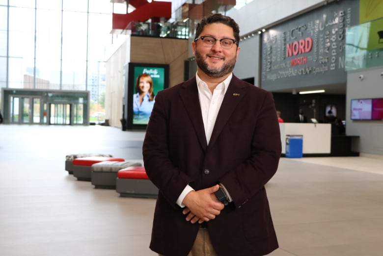 A man in a brown suit jacket and tan pants, wearing glasses, poses for a picture inside the atrium of the CBC/Radio-Canada building in Montreal. 