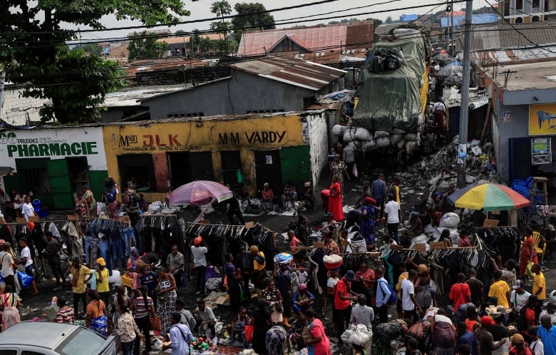 View from high up of colourful crowd, umbrellas, single-storey buildings with corrugated roofs, racks for clothing and jewelry in street.