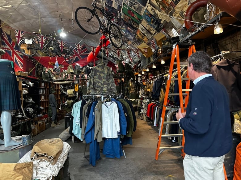 A man with dark hair wearing a blue shirt and white pants looks at a store filled with various shirts, posters, flags and other items. Several posters line the walls.