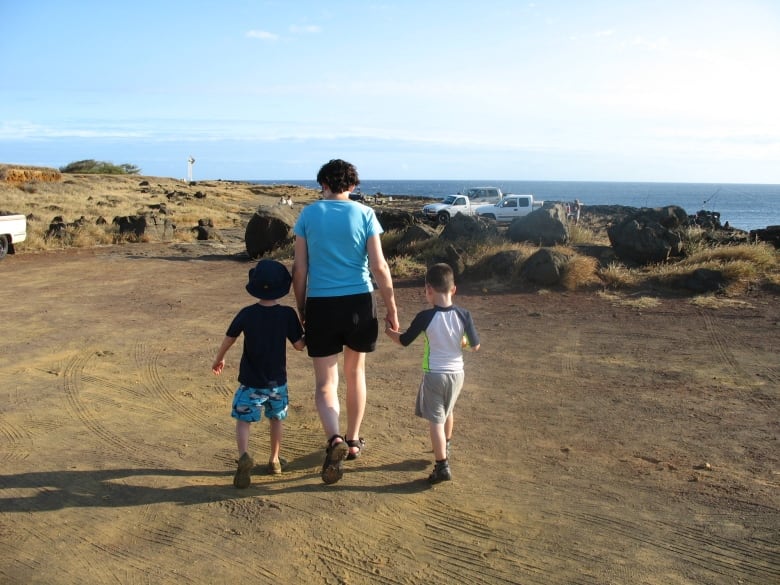 A woman holds hands with two elementary-school aged children as they walk toward a beach.