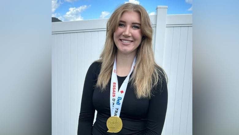 A young woman wearing a gold medal smiles as she stands outside.