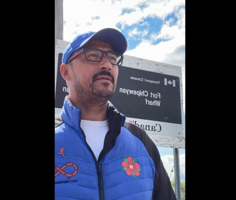 A guy stands in front of a sign. He is wearing a puffer vest and hat. 