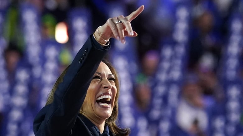 A woman in a dark suit laughs on a stadium stage and points. 