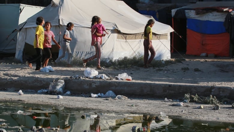 Several children are shown from a distance walking on a dirt road near tents. In the foreground, a large puddle.