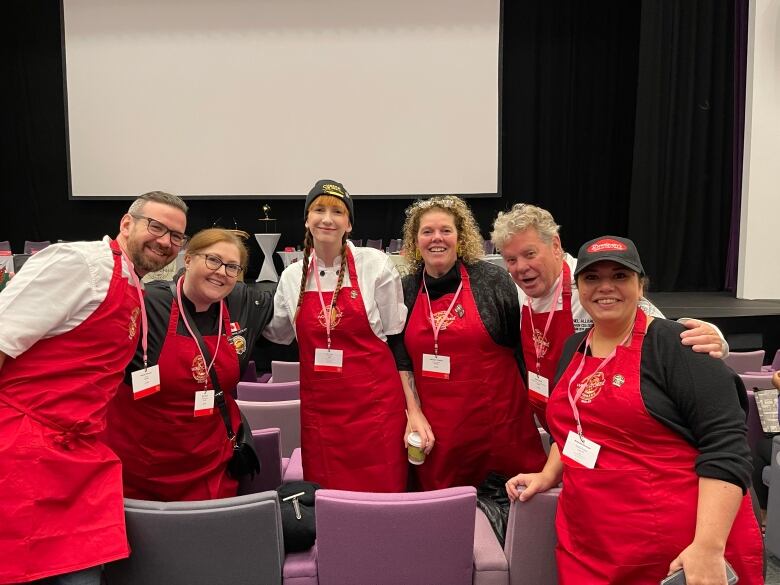 A panel of six cheese judges stand in red aprons.