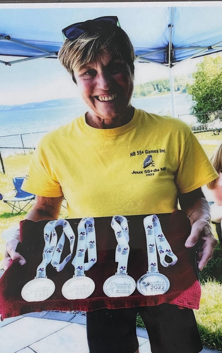 Woman with short hair with sunglasses on top of her head wearing yellow t-shirt holding up four medals to the camera and smiling.